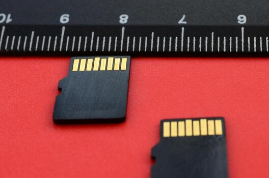 Two small micro SD memory cards lie on a red background next to a black ruler. A small and compact data and information store photo