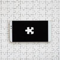 A modern big smartphone with several puzzle elements on the touch screen lies on a white jigsaw puzzle in an assembled state photo
