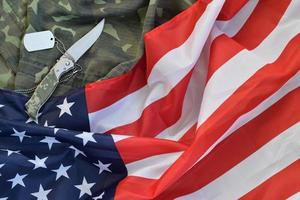 Army dog tag token and knife lies on Old Camouflage uniform and folded United States Flag photo