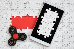 A modern big smartphone with a touch screen and a spinner lie on a white jigsaw puzzle in an assembled state with missing elements photo