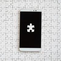 A modern big smartphone with several puzzle elements on the touch screen lies on a white jigsaw puzzle in an assembled state photo