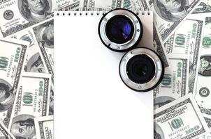 dos lentes fotográficos y un cuaderno blanco se encuentran en el fondo de muchos billetes de dólar. espacio para texto foto