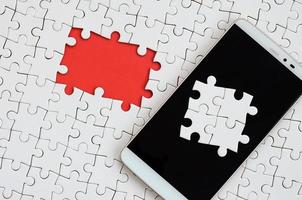 A modern big smartphone with several puzzle elements on the touch screen lies on a white jigsaw puzzle in an assembled state with missing elements photo