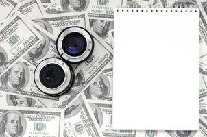 Two photographic lenses and white notebook lie on the background of a lot of dollar bills. Space for text photo