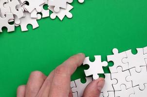 The hand folds a white jigsaw puzzle and a pile of uncombed puzzle pieces lies against the background of the green surface. Texture photo with space for text