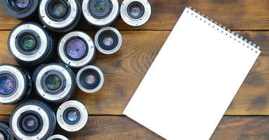 Several photographic lenses and white notebook lie on a brown wooden background. Space for text photo