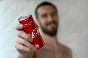 KHARKOV. UKRAINE - MAY 2, 2019 Smiling man gives non-alcoholic Coca-Cola aluminium tin can in garage interior photo