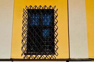 A large rectangular window of a yellow stone building closed by a large black iron grate. The background photo