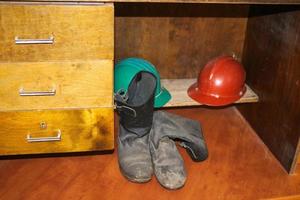 casco de seguridad de plástico naranja y amarillo para el trabajador. casco protector para proteger la cabeza de las personas que operan en condiciones peligrosas y botas negras de goma en la fábrica debajo de la mesa foto