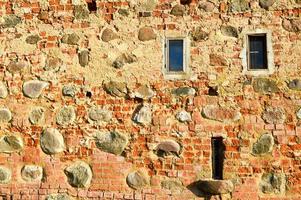 Texture with small narrow windows on an old ancient stone cracked dilapidated brick wall of red brick with large boulders. The background photo