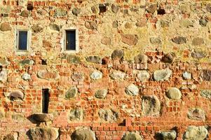 Texture with small narrow windows on an old ancient stone cracked dilapidated brick wall of red brick with large boulders. The background photo