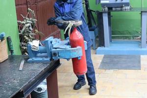 A man working on a large metal industrial vice with a huge gas wrench unscrews a nut on a red fire extinguisher cylinder in a factory workshop photo