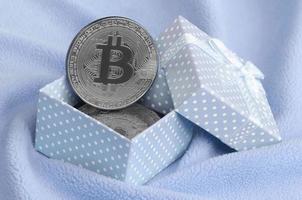 The silver bitcoin lies in a small blue gift box with a small bow on a blanket made of soft and fluffy light blue fleece fabric with a large number of relief folds photo