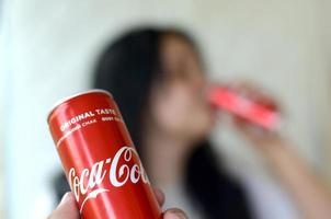 Jarkov. ucrania - 2 de mayo de 2019 mujer morena bebe coca-cola en el interior del garaje y la mano masculina presenta una lata de cola en foco foto