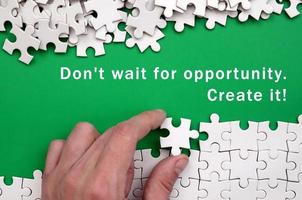 Don't wait for opportunity. Create it. The hand folds a white jigsaw puzzle and a pile of uncombed puzzle pieces lies against the background of the green surface photo