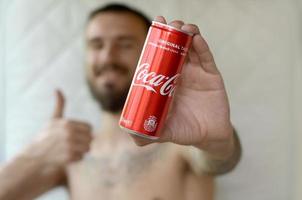 Jarkov. ucrania - 2 de mayo de 2019 un hombre sonriente presenta una lata de aluminio de coca-cola sin alcohol con un símbolo similar en el interior del garaje foto