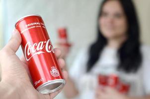 KHARKOV. UKRAINE - MAY 2, 2019 Happy woman holding few Coca-Cola tin cans in garage interior and male hand with one can in foreground photo