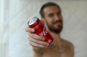 KHARKOV. UKRAINE - MAY 2, 2019 Smiling man gives non-alcoholic Coca-Cola aluminium tin can in garage interior photo