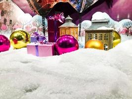 round, glass, multi-colored balls lie on the white snow. pink and gold ball lie on the background of small houses. new year decoration, christmas eve photo