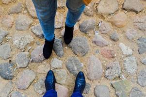 Male and female legs in leather shoes, boots on a stone road of large cobble stones opposite each other. The background photo