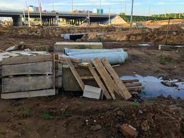 sitio de construcción, charcos de arena y lodo. hay estructuras de madera en el sitio. contra el fondo del puente, cruce de carreteras. tuberías y materiales de construcción yacen en el suelo foto