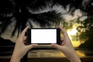 Hand holding smartphone with white blank screen over blurred silhouette of coconut trees on the beach background. photo