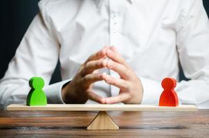 A man with hands in the lock and looks at the rival red and green figures on a scales. The concept of conflict resolution and the search for a compromise in the dispute. Weighted decision photo