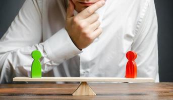 Man holds hand on chin looks at the rival red and green figures on a scales. Weighted decision. The concept of conflict resolution and the search for a compromise in the dispute. photo