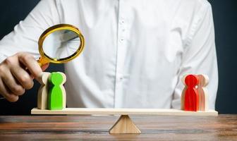 A man with a magnifying glass is looking at the rival red and green figures groups on scales. conflict resolution and the search for a compromise in the dispute. Research argument of each side photo