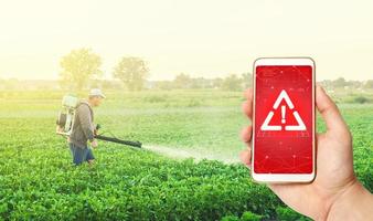 Phone in hand displaying a warning against background of a farmer with a mist sprayer fogger on a farm field. Use of dangerous chemicals for the treatment of plants and crops. Potential health hazard. photo