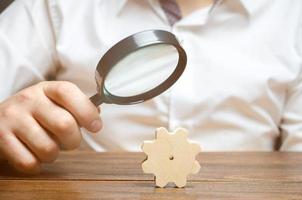 Businessman examines a wooden gear through a magnifying glass. study and analysis of business processes and subjects. A cog from a big business machine. Every little thing is important. Research photo