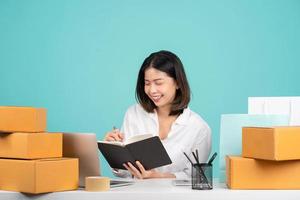 Smiling Asian businesswoman in a casual shirt sits at a desk with a brown cardboard box. She is taking notes on a conversation, isolated on a green background. photo