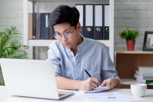 Young Asian businessman sitting at a desk is writing notes in a notebook using a laptop computer at home, working at home. photo