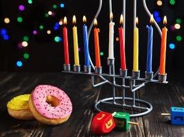 Jewish holiday Hanukkah background. A traditional dish is sweet donuts. Hanukkah table setting candlestick with candles and spinning tops on black background. Lighting Chanukah candles. Copy space photo