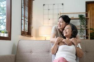 Portrait happy adult daughter and older mother hugging and holding hands, sitting on couch at home, young woman and mature mum or grandmother looking at camera, two generations family photo