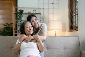 Portrait happy adult daughter and older mother hugging and holding hands, sitting on couch at home, young woman and mature mum or grandmother looking at camera, two generations family photo