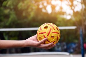 joven jugador de sepak takraw masculino del sudeste asiático usando su mano derecha para sostener la pelota antes de enviar a otro jugador a patear, sepak takraw al aire libre jugando después de la escuela, enfoque suave y selectivo en la pelota. foto