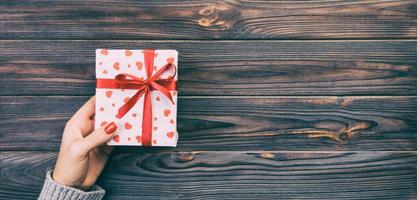 Woman hands give wrapped valentine or other holiday handmade present in paper with red ribbon. Present box, red heart decoration of gift on wooden table, top view with copy space Toned web banner photo