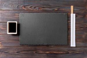 Empty rectangular black slate plate with chopsticks for sushi and soy sauce on wooden background. Top view with copy space photo