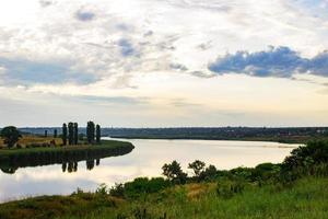 Summer evening dawn sky clouds reflected on a lake of water on the coast with green grass and forest trees photo