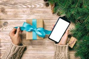 female hands hold wrapping christmas gift box and reading massage on phone with blue ribbon on brown wooden vintage table. Top view photo