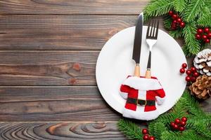 Holiday composition of plate and flatware decorated with Santa clothes on wooden background. Top view of Christmas decorations with empty space for your design. Festive time concept photo