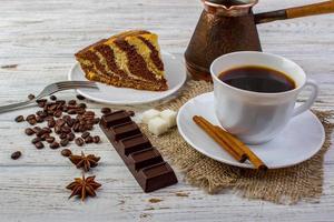 A cup of coffee on sacking with tasty chocolate cake on a plate. Cinnamon sticks, sugar, coffee beans, a chocolate bar, anice on bright wooden table photo
