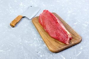 pedazo grande de carne fresca en una tabla de cortar de madera con hacha de cocina. vista superior sobre fondo de hormigón gris foto