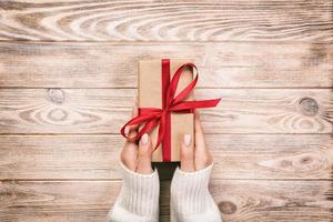 Woman hands give wrapped valentine or other holiday handmade present in paper with red ribbon. Present box, decoration of gift on white wooden rustic vintage table, top view with copy space photo