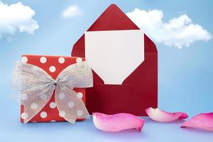 Burgundy letter with paper for text stands. Red polka dot gift box with silver bow and rose petals on blue background with clouds. Holiday, invitation. Birthday, March 8, Saint Valentin photo