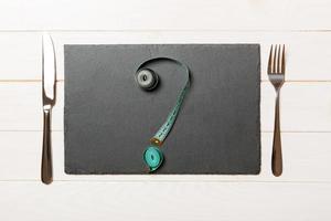 Top view of slate plate, fork and measure tape in form of question mark on wooden background. Overeating concept and copy space photo