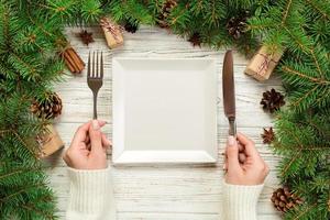 top view girl holds fork and knife in hand and is ready to eat. Empty white square plate on wooden christmas background. holiday dinner dish concept with new year decor photo