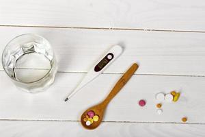 table spoon with tablets, thermometer and glass of water on white wooden table. Medical concept photo