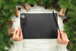 top view girl holds fork and knife in hand and is ready to eat. Empty black slate rectangular plate on wooden christmas background. holiday dinner dish concept with new year decor photo